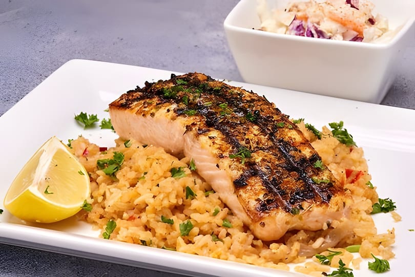 Grilled salmon with rice pilaf and a lemon wedge, served with a side of coleslaw.