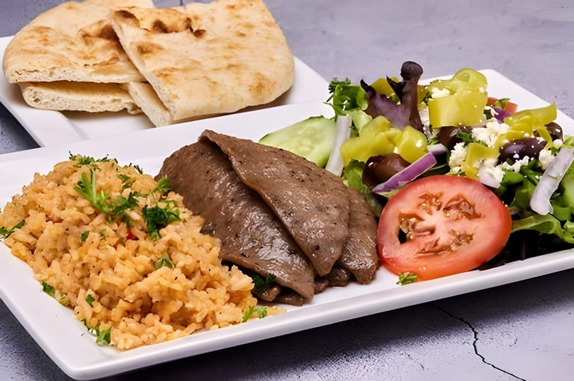 A gyro platter with sliced meat, rice, Greek salad, and pita bread on a white plate.