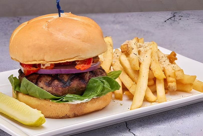 Gourmet burger with lettuce, onion, tomato, and a side of seasoned fries and a pickle spear.