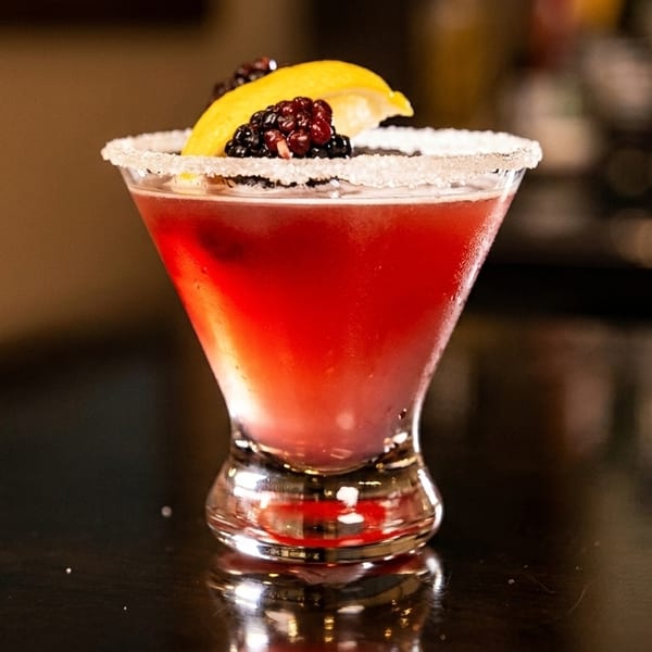 A vibrant red cocktail in a sugar-rimmed martini glass, garnished with blackberries and a lemon slice.